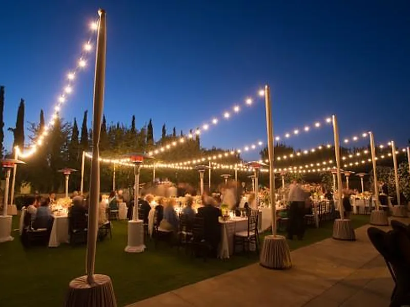 Warm market string lights strung over guests at round tables during an outdoor evening party