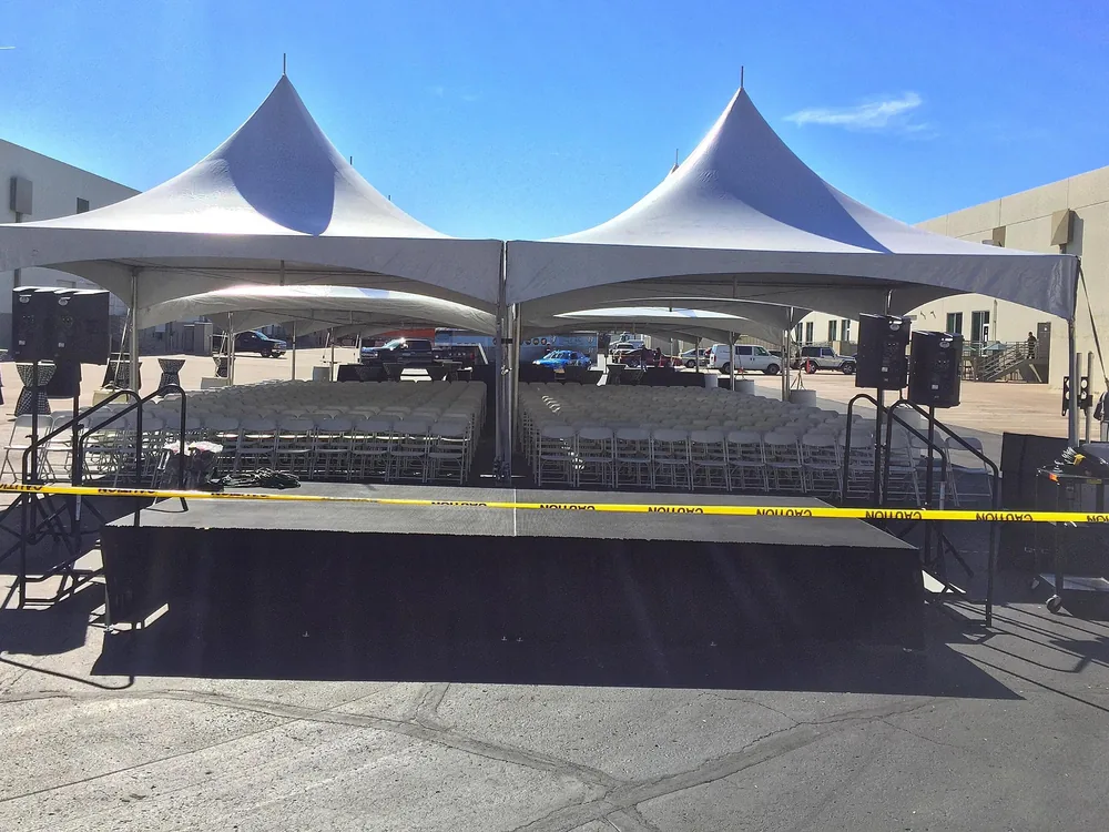 Black portable stage set up outdoors with speakers and stair rails on each side, in front of white peaked tents filled with rows of folding chairs